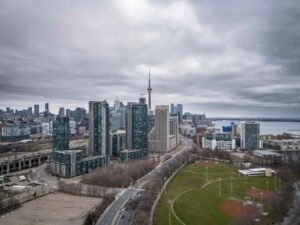Cityscape with tall buildings, central tower, road, sports field, water, and overcast sky.
