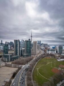 Urban skyline with tower, tall buildings, roadway traffic, green park, and cloudy sky over water.