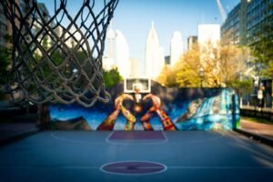 Basketball hoop on street court, urban mural and city buildings under clear sky in background.