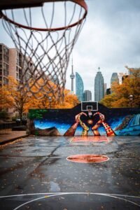 Wet basketball court with a mural of abstract patterns, hands, urban buildings, and orange trees.