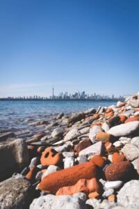 Rocky shoreline with stones, city skyline across water under a clear blue sky in the background.