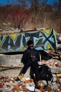 Person on concrete with phone, surrounded by bricks, graffiti wall, dried plants, black backpack.