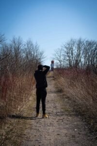Person on dirt path photographing lighthouse amid dry grass and trees under clear sky.