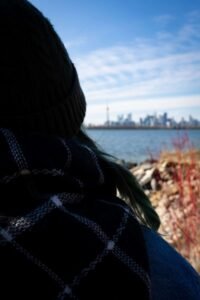 Person in beanie and plaid scarf by water, city skyline in distance, red plants nearby.