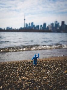 Small blue toy on pebbly beach, city skyline with tower under cloudy sky in background.