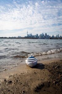 Toy cruise ship on sandy beach with city skyline and tower in background under cloudy sky.