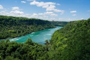 Wide turquoise river in lush forest, bordered by steep cliffs under a blue sky with clouds.