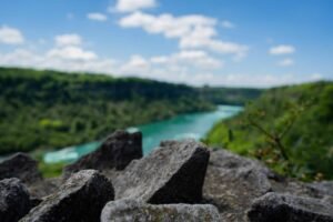 Sharp rocks in focus, river and green hills blurred in background under blue sky with clouds.