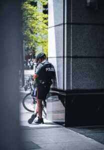 Police officer by a bicycle leaning on building corner, tree and people in background.