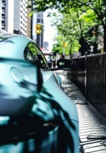 Sleek car on urban sidewalk, sunlight through trees, tall buildings, blurred traffic light.