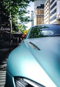 Metallic blue sports car on city street, shadows from trees, urban buildings reflected.