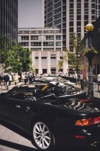 Black convertible parked on city street with crowd, buildings, trees, and floral lamp post.