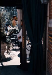 Person in white shirt and mask partially visible in doorway, with outdoor seating and plants.