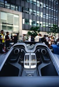 Rear of sleek sports car with vents, engine details; people on city street by office building.