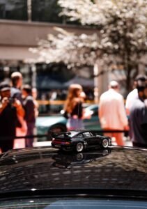 Miniature black car model on vehicle roof, outdoor setting with blurred figures and tree background.