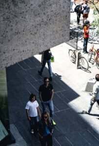 Street scene with pedestrians on a sunny day, bicycles parked nearby, shadows visible.