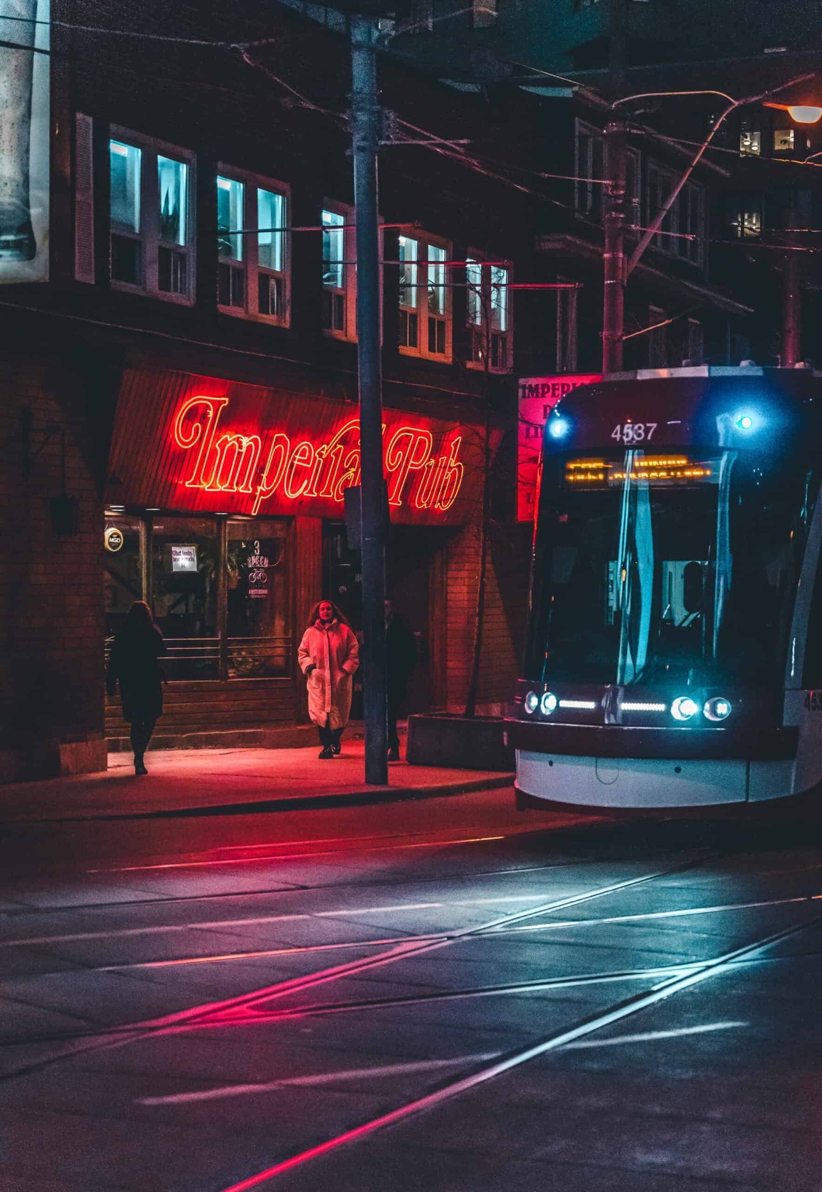 Streetcar at night with red pub sign, wet pavement, and pedestrians nearby.