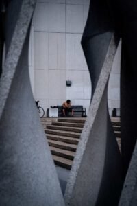 Person sitting on bench beside bicycle, urban setting with large stone structures.