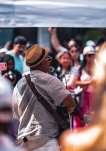 Musician in hat and sunglasses plays guitar and sings into microphone at outdoor event.