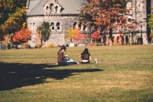 People sitting on grass near a historic stone building with fall trees in the background.